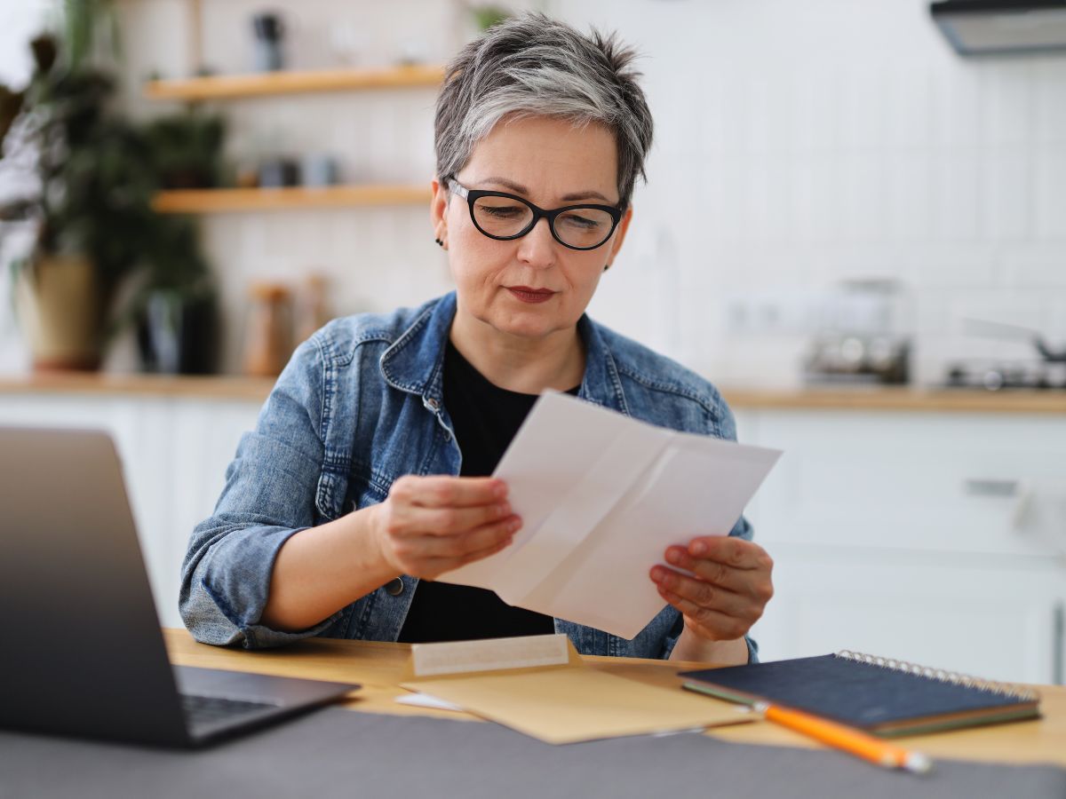 A woman wearing glasses sits at a desk with a laptop and notebook, reading a document she has just opened from an envelope.