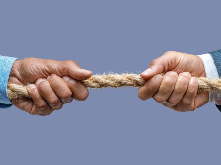 Two hands in business attire pull a fraying rope from opposite ends against a gray-blue background, illustrating a tug of war between two opposing forces.