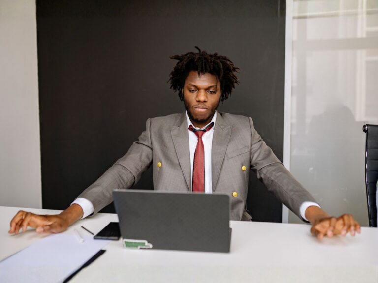 A man in a gray suit and red tie sits at a desk with a laptop, looking down with a serious and focused expression.
